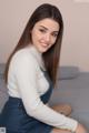 A woman sitting on a bed wearing a white sweater and blue overalls.